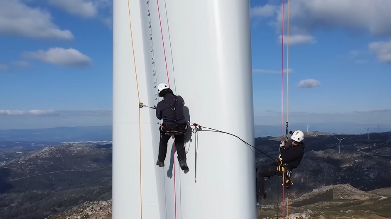 Two Wind Turbine Technicians Are Rappelling Down a Wind Turbine in A Mountainous Area with Other Turbines Visible in The Background