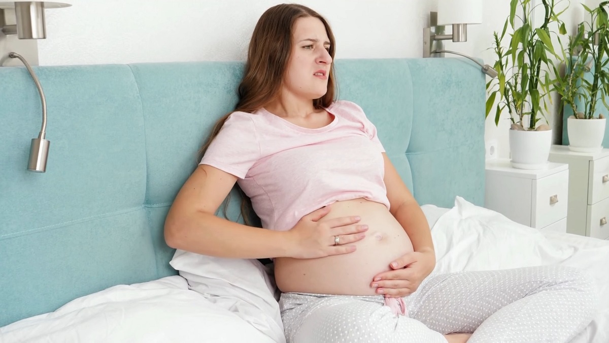 A pregnant woman sits on a bed, holding her belly with a distressed expression